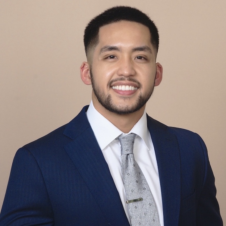 man with dark hair and beard wearing blue suit and light gray tie in front of tan background