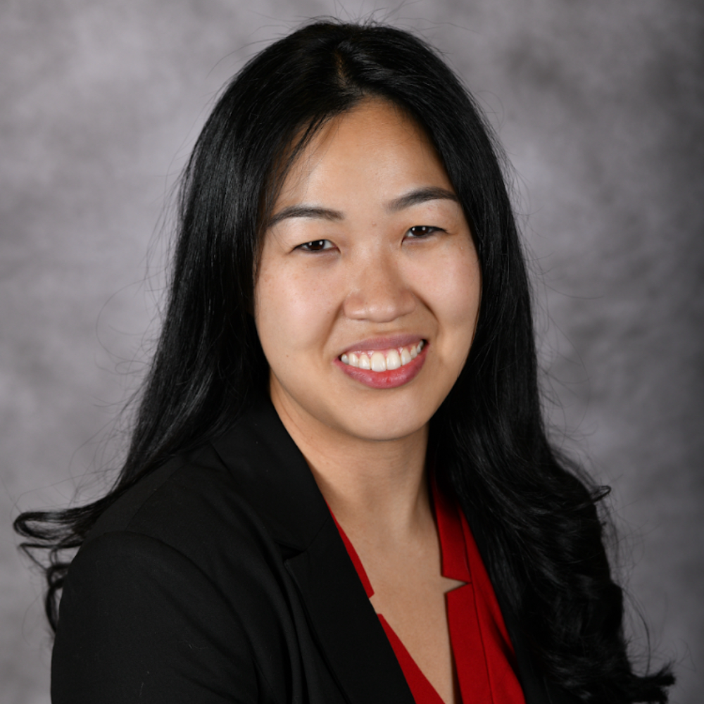 asian woman with long dark hair wearing black blazer and red top with gray background
