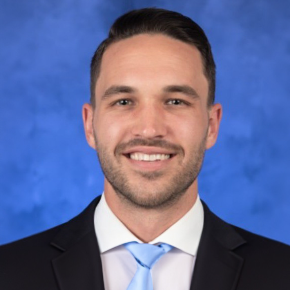 man with dark hair and facial hair wearing black suit in front of blue background