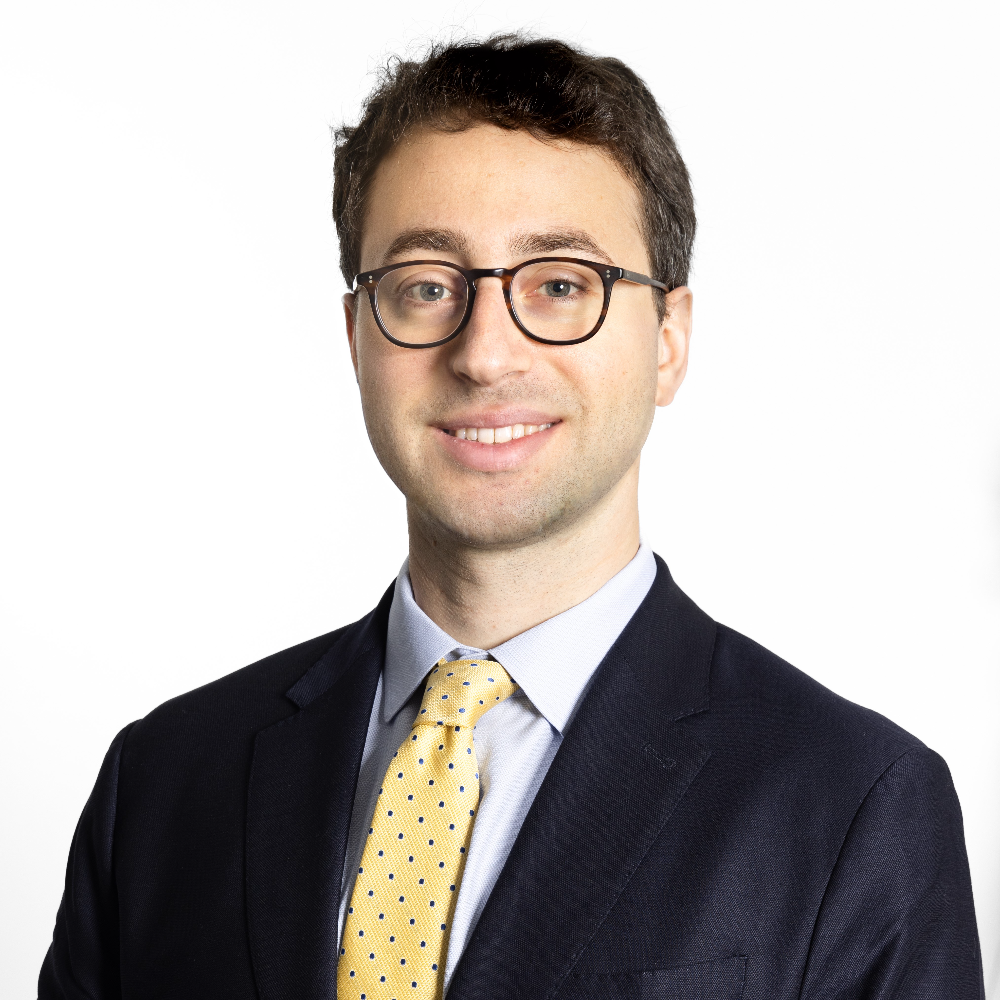 man with short dark hair wearing glasses and dark suit with yellow tie