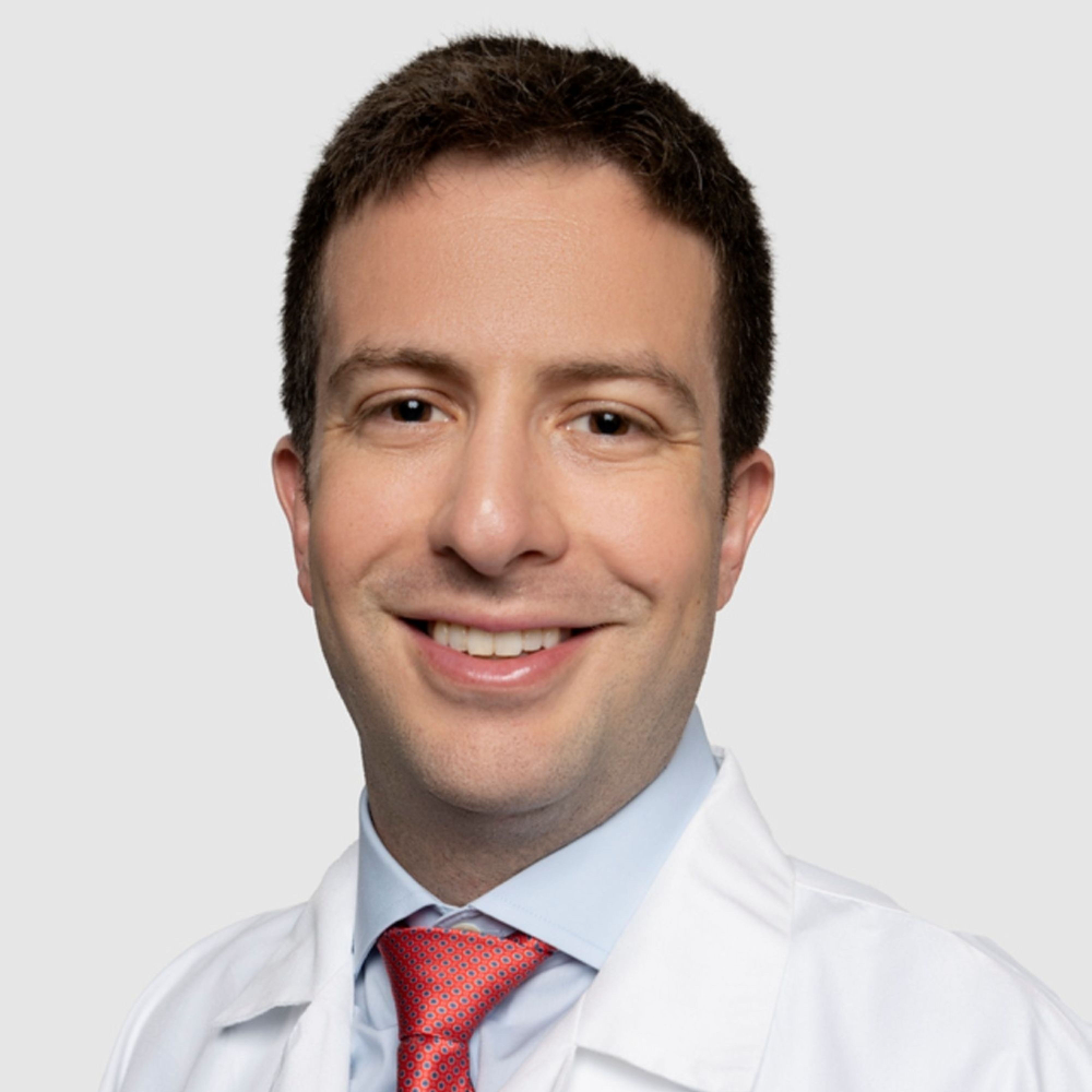man with short dark hair wearing white coat and light blue shirt and red tie