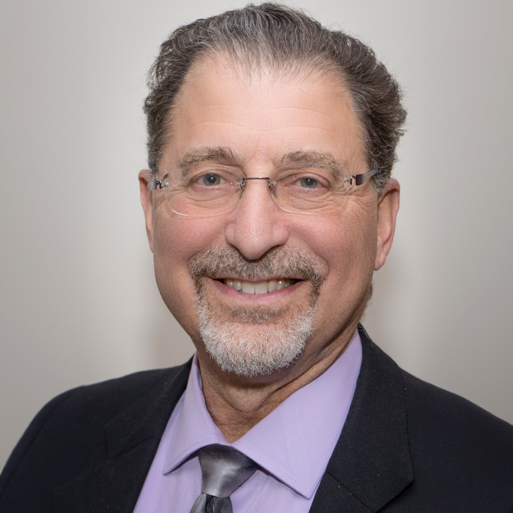 man with dark hair and beard wearing glasses wearing dark suit and lavender shirt