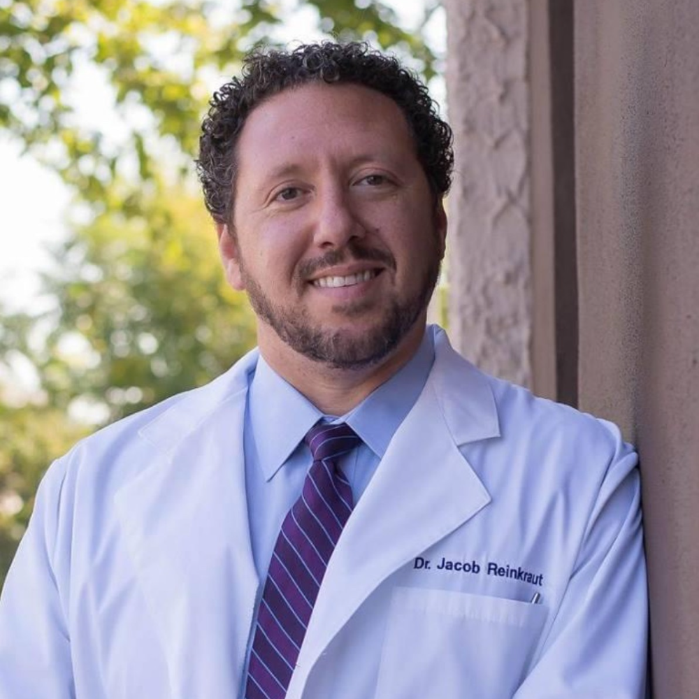 man with dark hair and beard wearing white coat standing outside