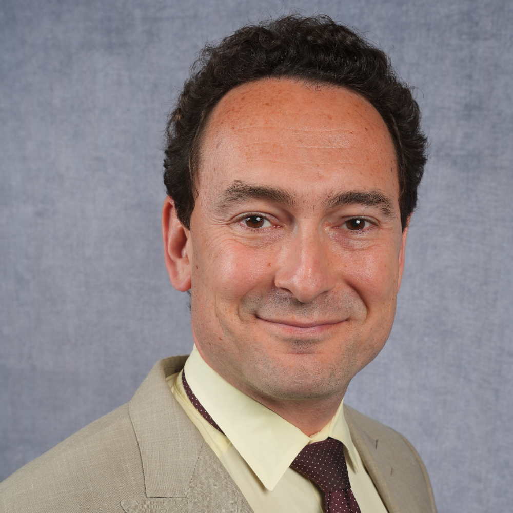 man with short brown hair earing a tan suit jacket with yellow shirt and tie in front of a gray background