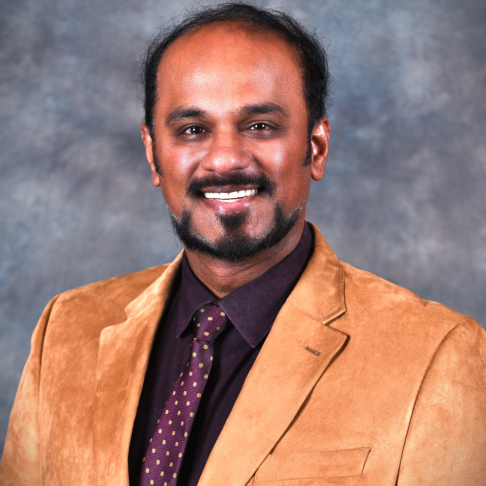 man with dark hair and beard wearing tan sports jacket and dark shirt