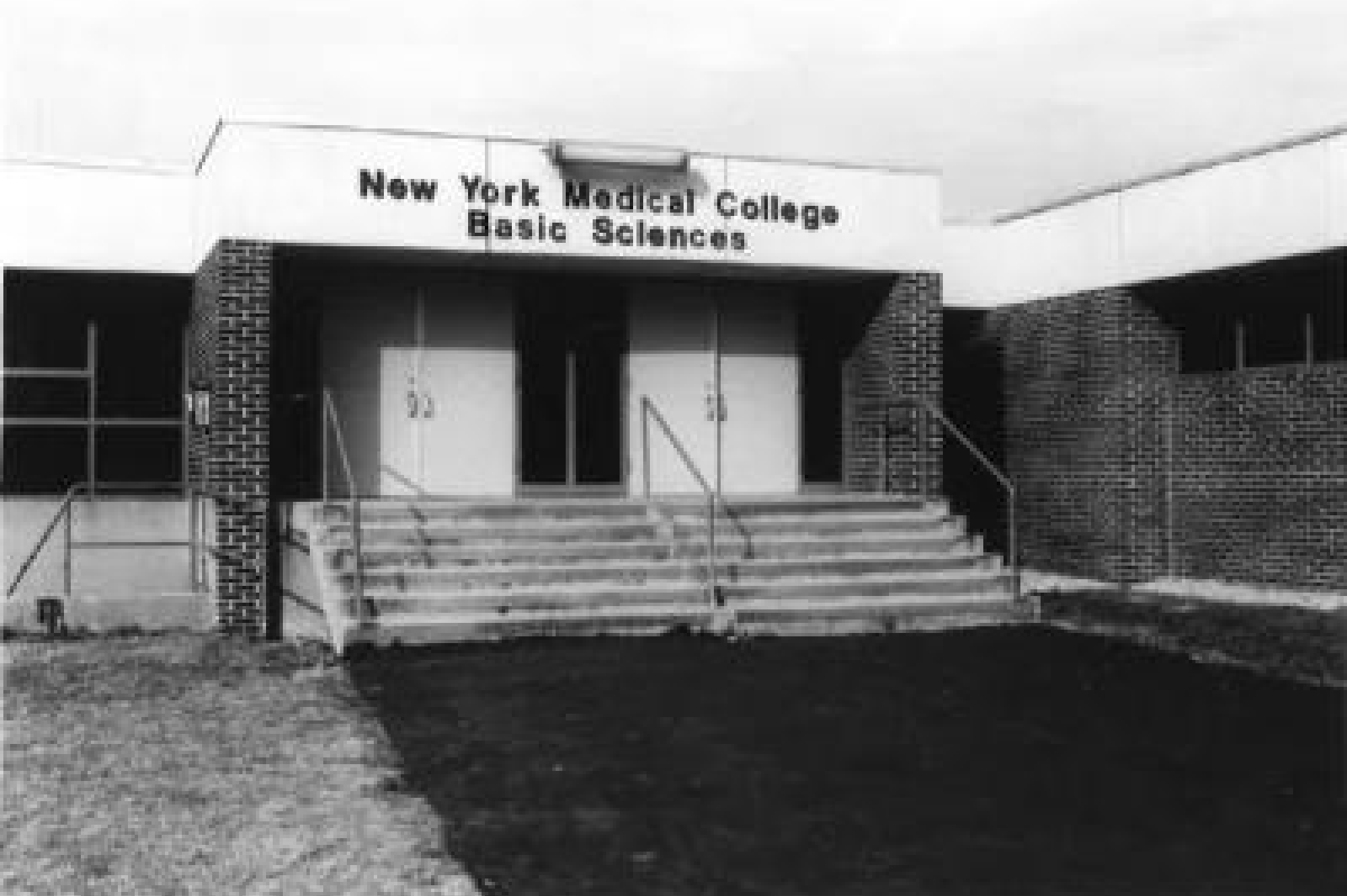 Brick building with staircase in front and white trim at top with sign that says New York Medical College Basic Sciences Building. Staircase leads up to building