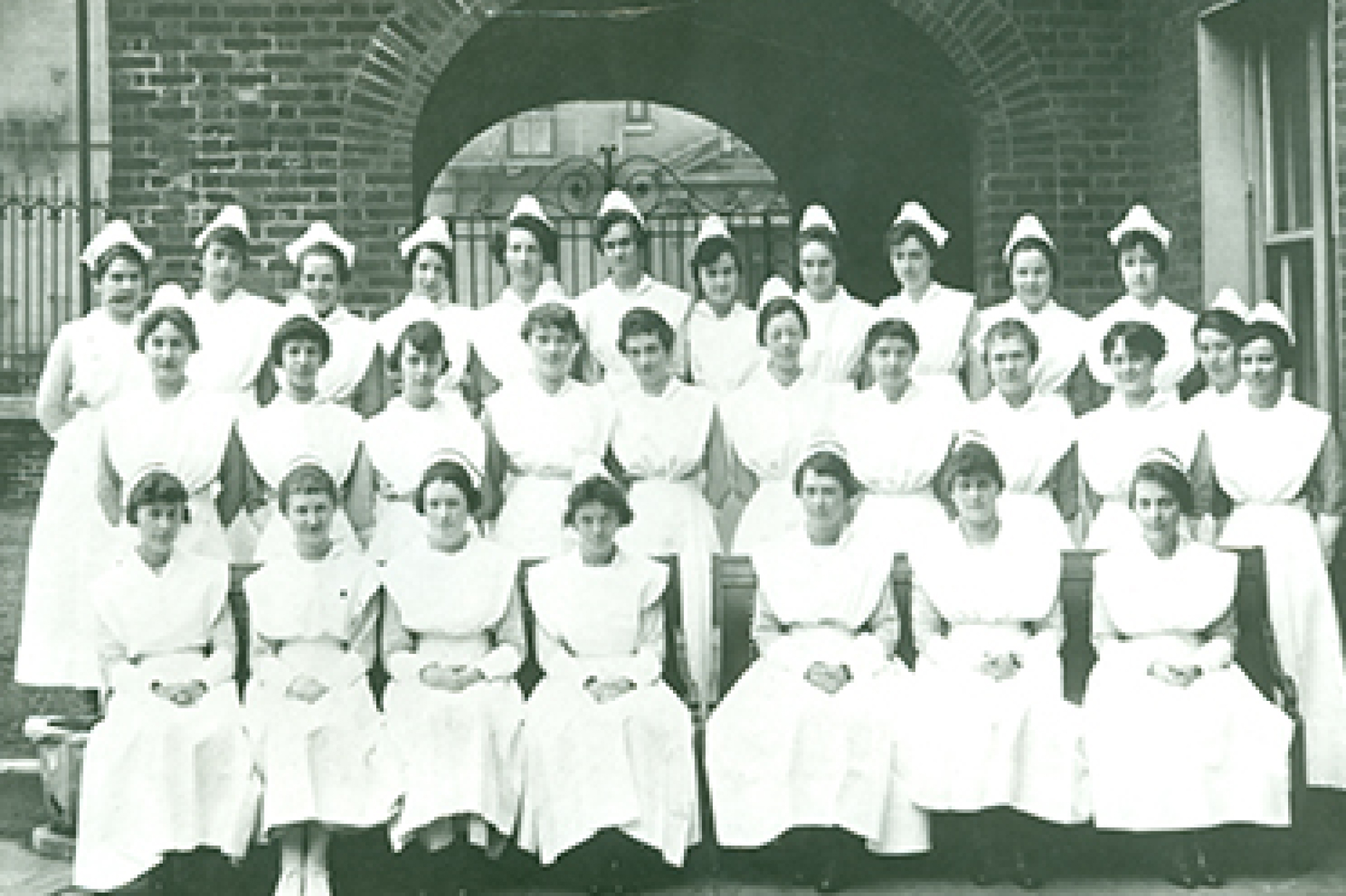 Three rows of female nursing students posing in their white uniforms in front of a brick archway