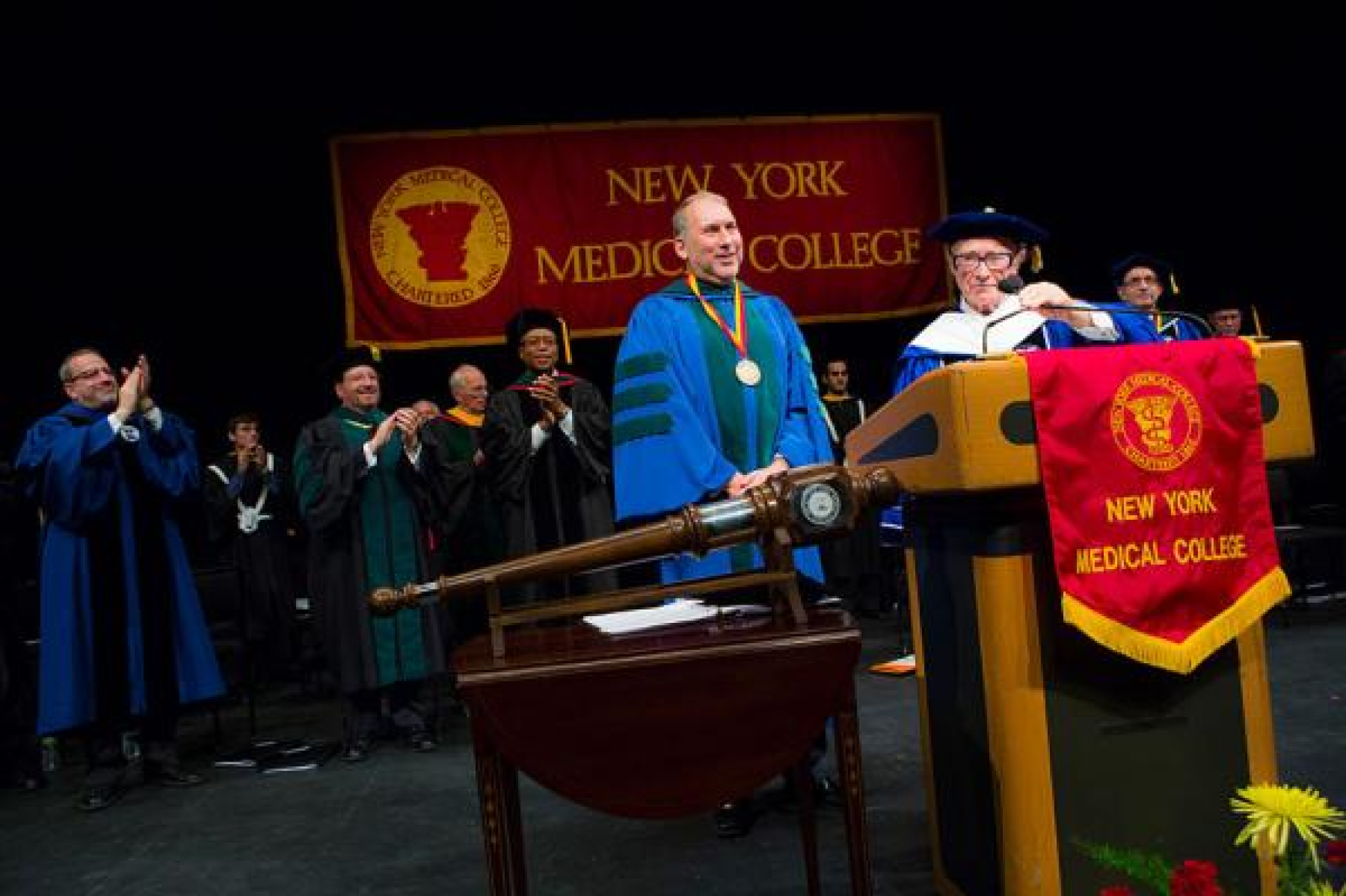 Dr. Halperin on stage at installation ceremony, wearing a blue robe, with NYMC and Touro administration clapping in background