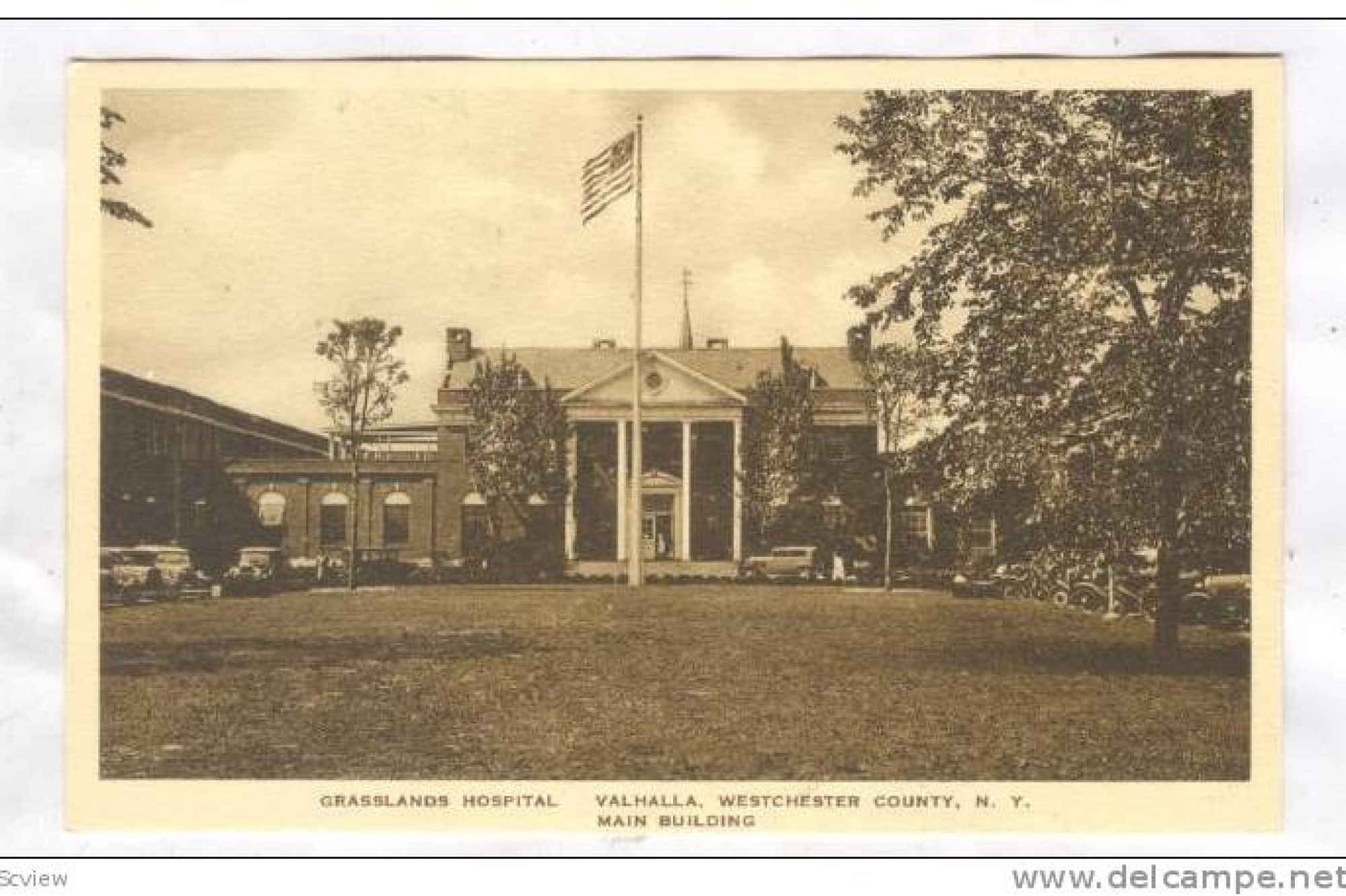 Old photo of the Grasslands Hospital, a multi-story building with large pillars and a large grassy area in front with a flag pole flying the American flag