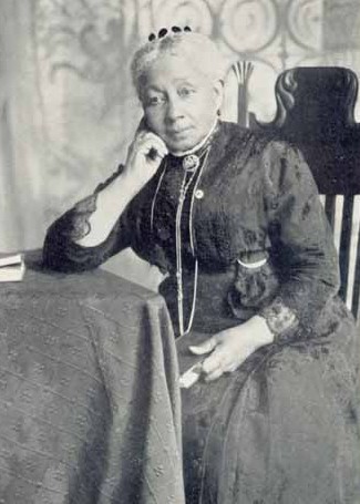 Black and white photograph of an elderly Susan McKinney Steward. She is a Black woman with white hair wearing a dark lace dress, a small headpiece, and several thin chain necklaces.