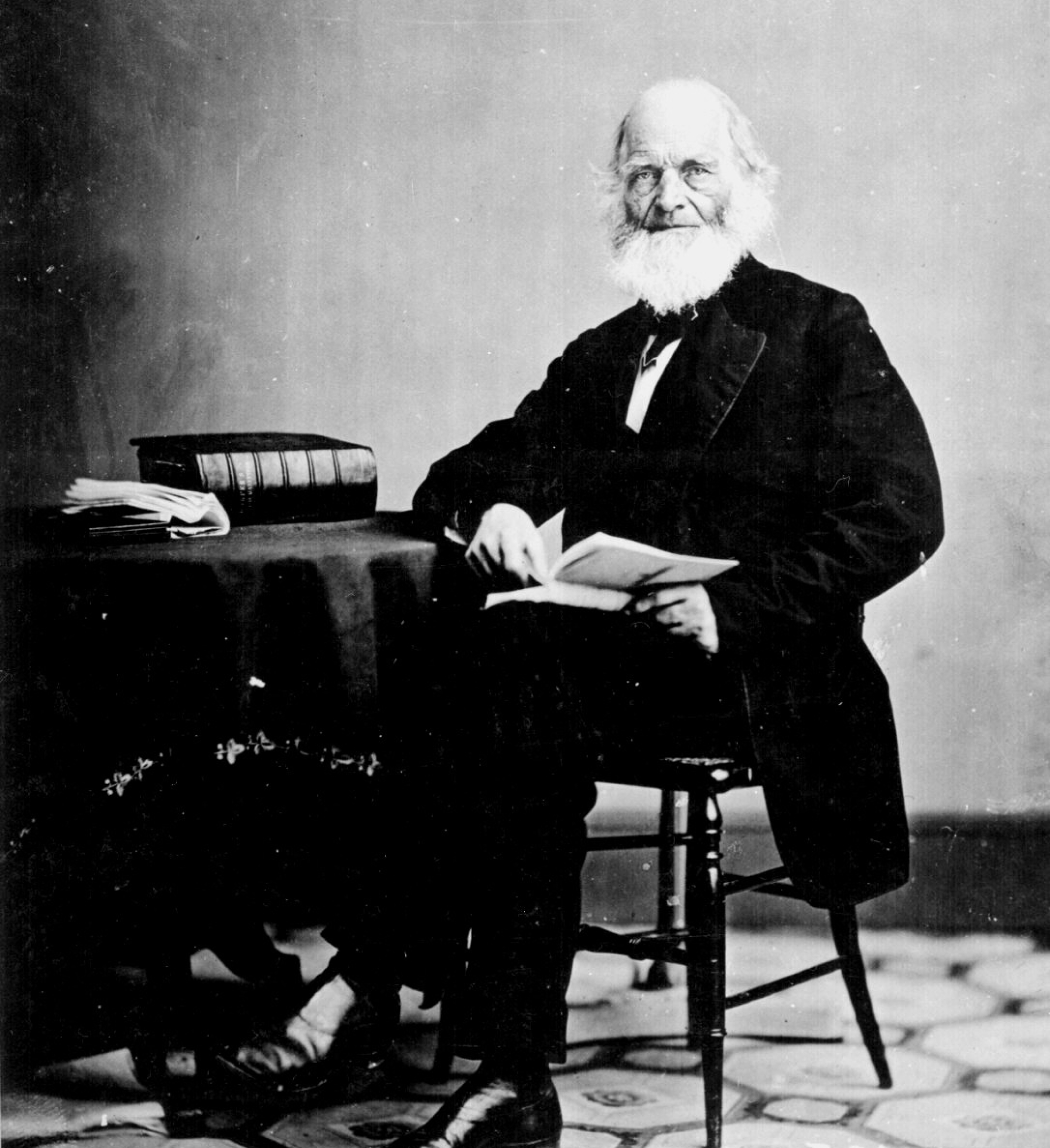 An elderly William Bryant with a white beard, wrinkled skin, and bald head, wearing a suit while sitting at desk with a book